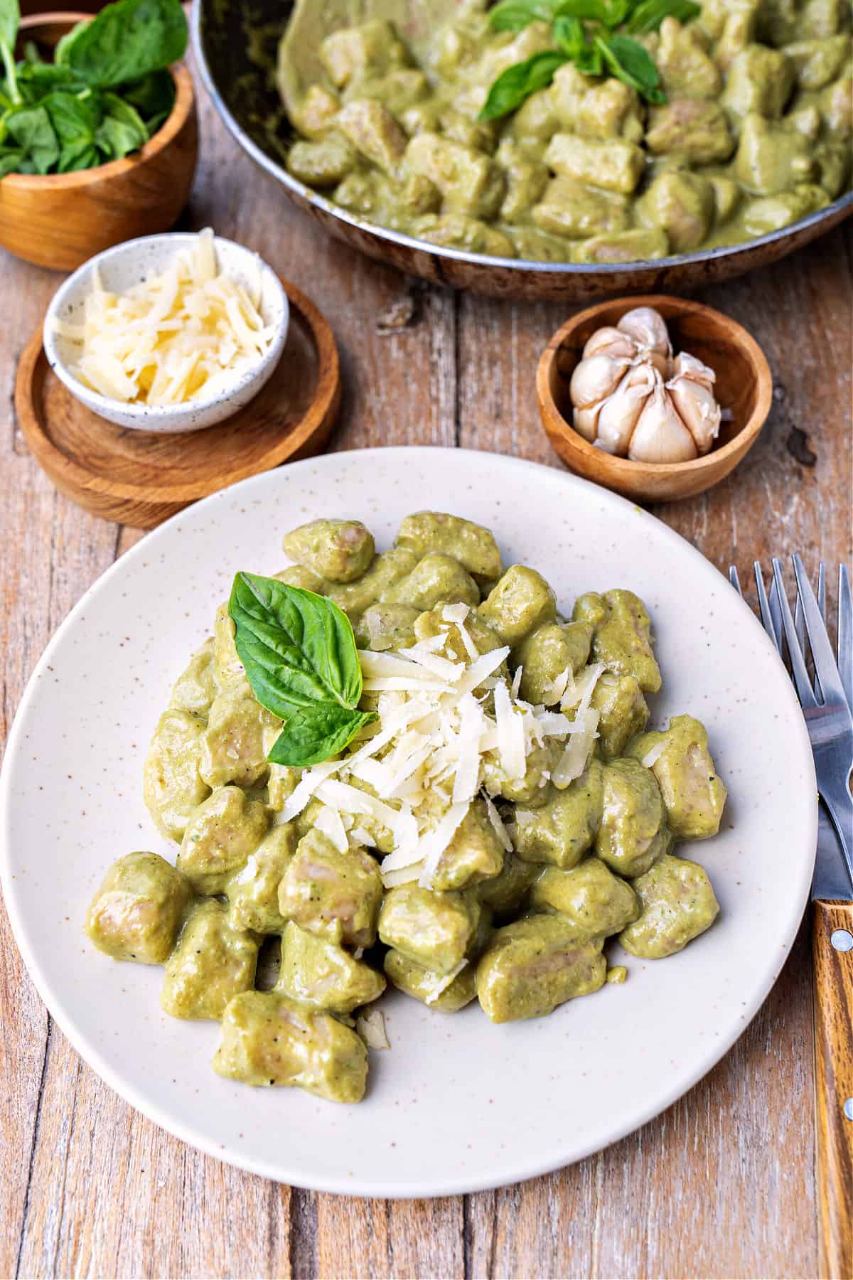 Wooden surface with white plate of pesto gnocchi. Grated cheese and garlic in bowls.