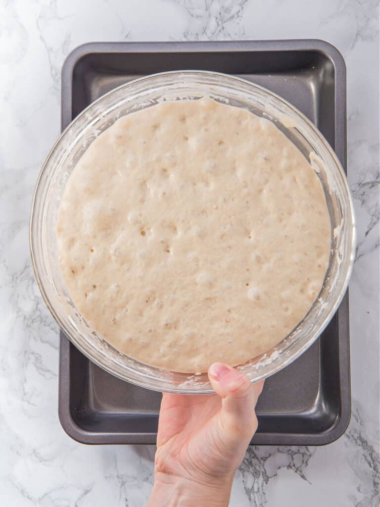 Risen focaccia dough in a glass bowl over baking pan on a marbled surface. Top view.