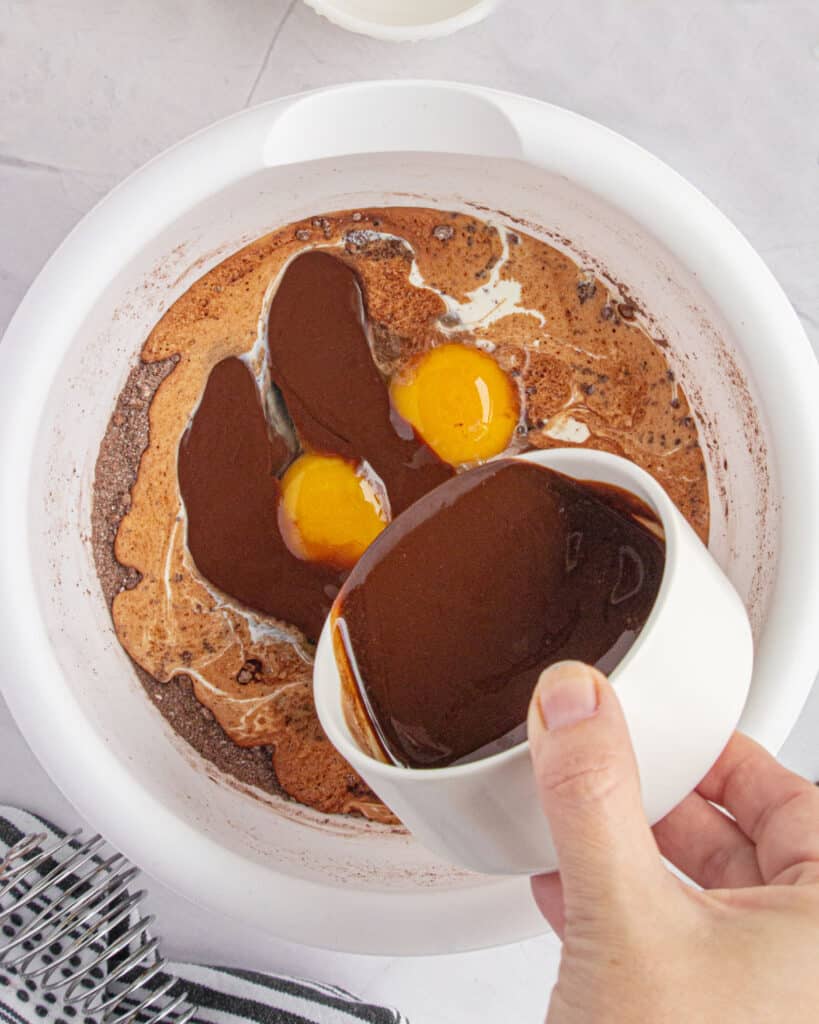 Pouring melted chocolate onto white bowl with eggs and dry ingredients.