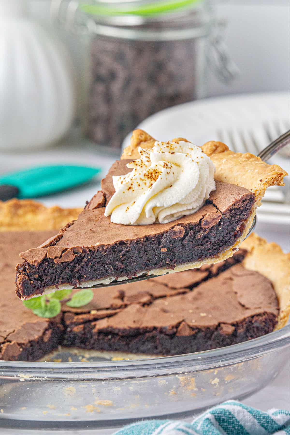 Slice of chocolate chess pie with cream dollop being lifted over whole pie in glass plate.
