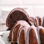 Close up of glazed chocolate bundt cake slice lifted from whole cake. White background.