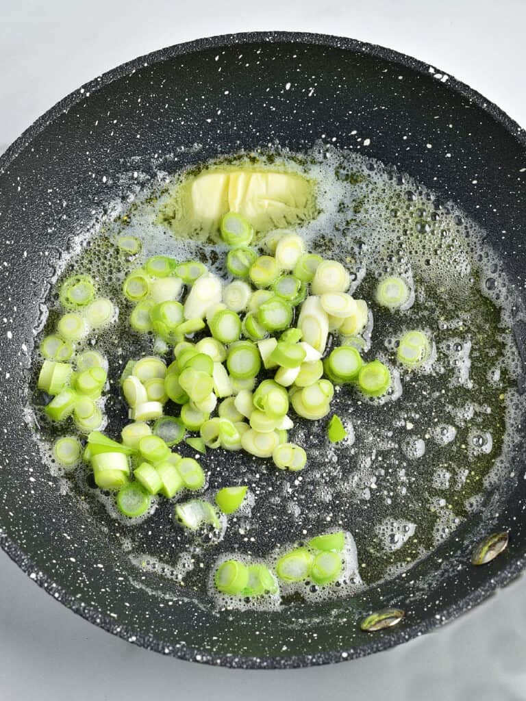 Chopped leeks and spring onion cooking with butter in a dark skillet.
