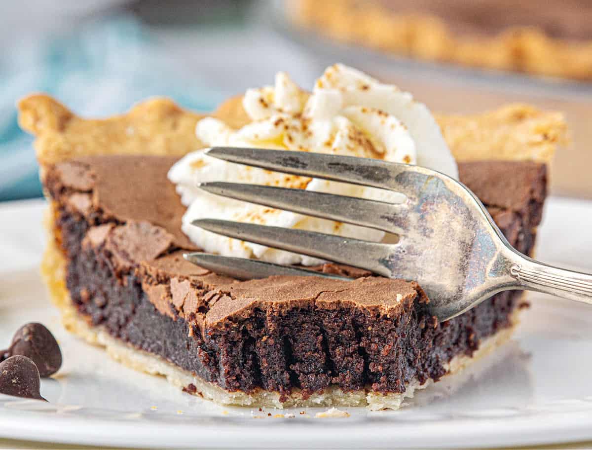 Forking into bitten slice of chocolate chess pie with cream dollop on a white plate.