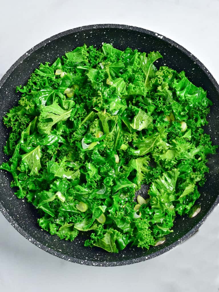 Kale added to dark skillet with leeks. Top view. Light gray background.
