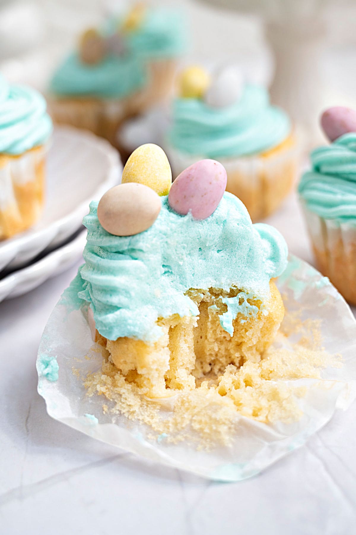 Bitten Easter mini egg cupcake in opened white paper liner. More cupcakes in the white background.