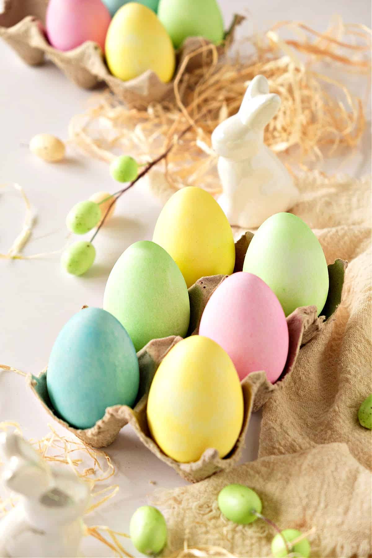 Egg cartoon with colored eggs. Mini eggs, shredded paper and bunny on a whitish background.