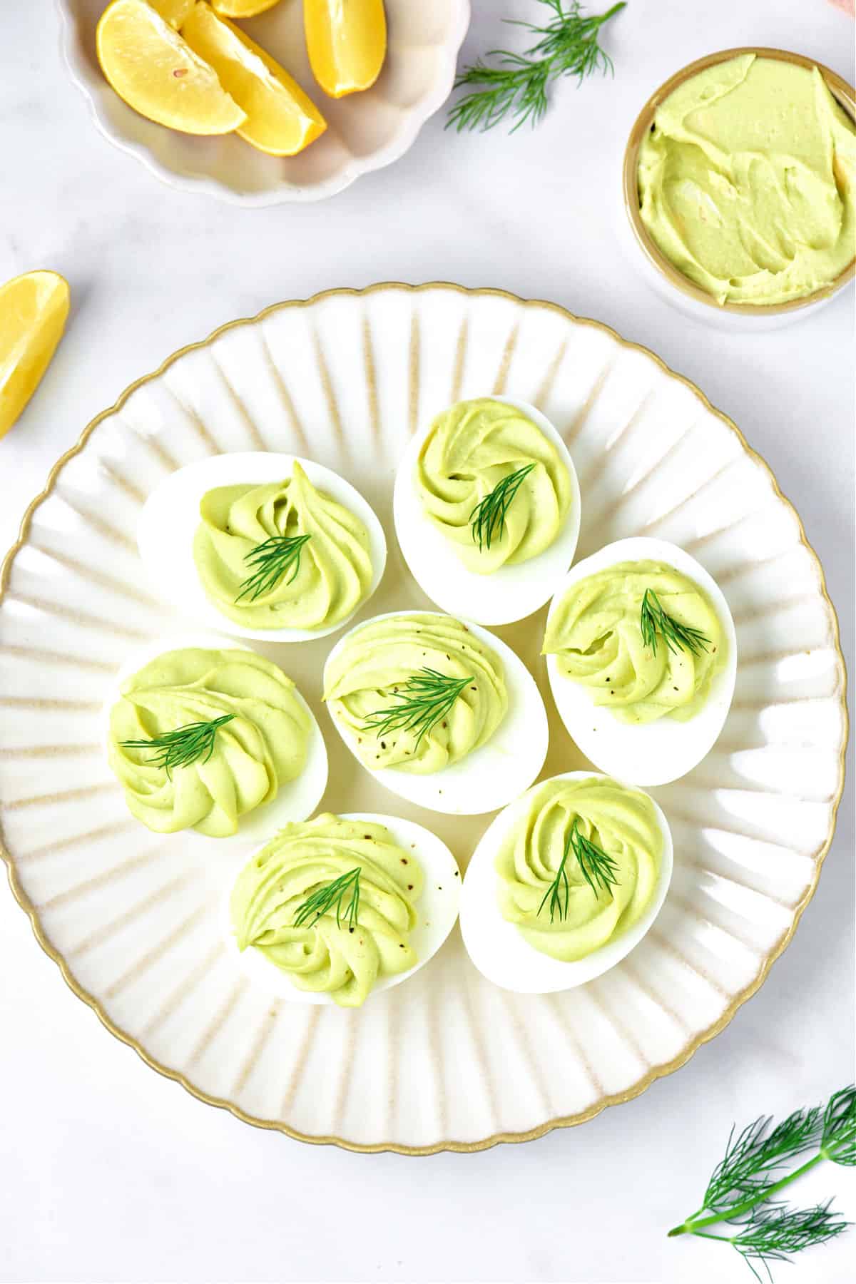 Several avocado deviled eggs with dill on a scalloped white beige plate. Lemon wedges, white background. Top view.
