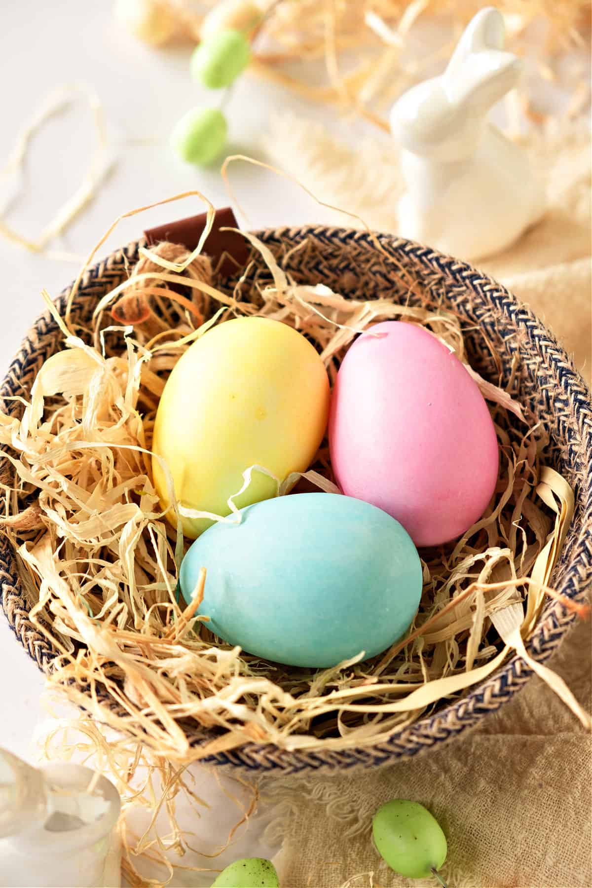Brown woven basket with colored eggs and shredded kraft paper. Ceramic bunny in the background.