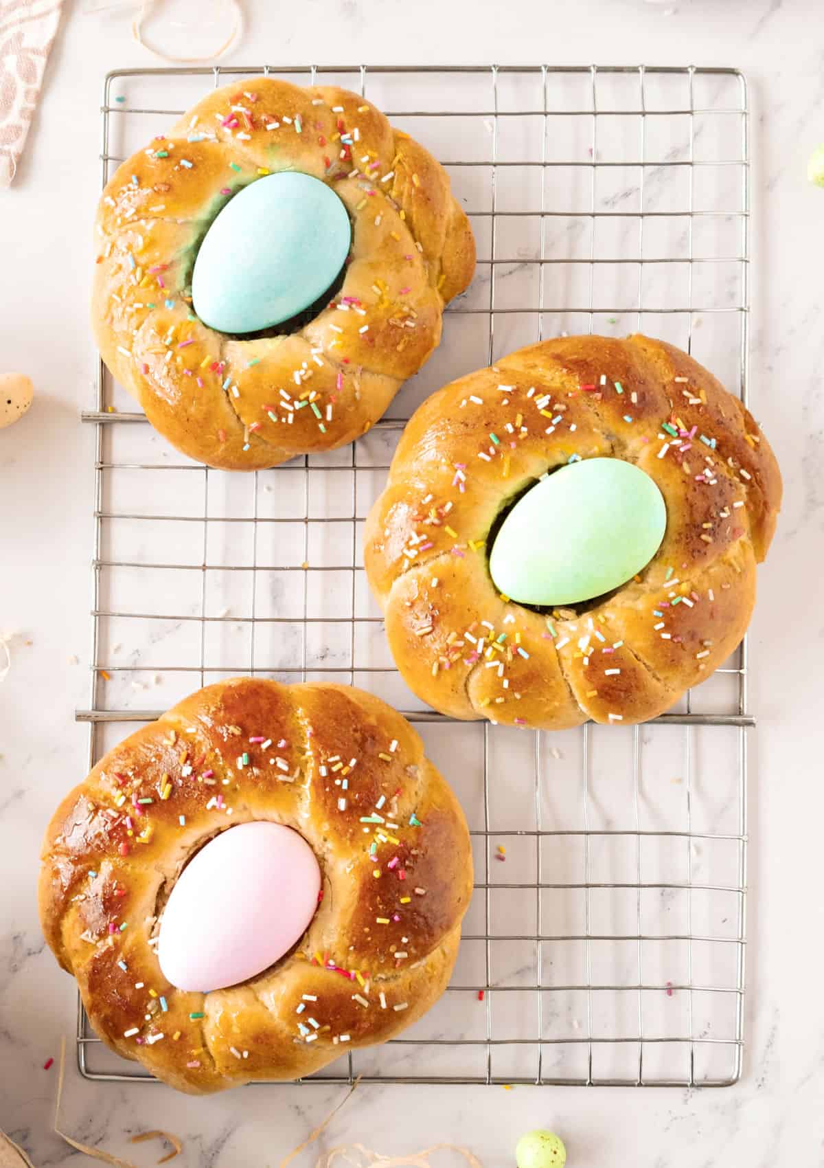 Three Easter braids with dyed eggs on a wire rack. White marble background, top view.