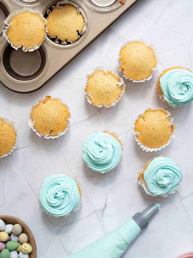 Frosted and plain cupcakes on a marbled surface. Piping bag with aqua frosting. Metal pan.