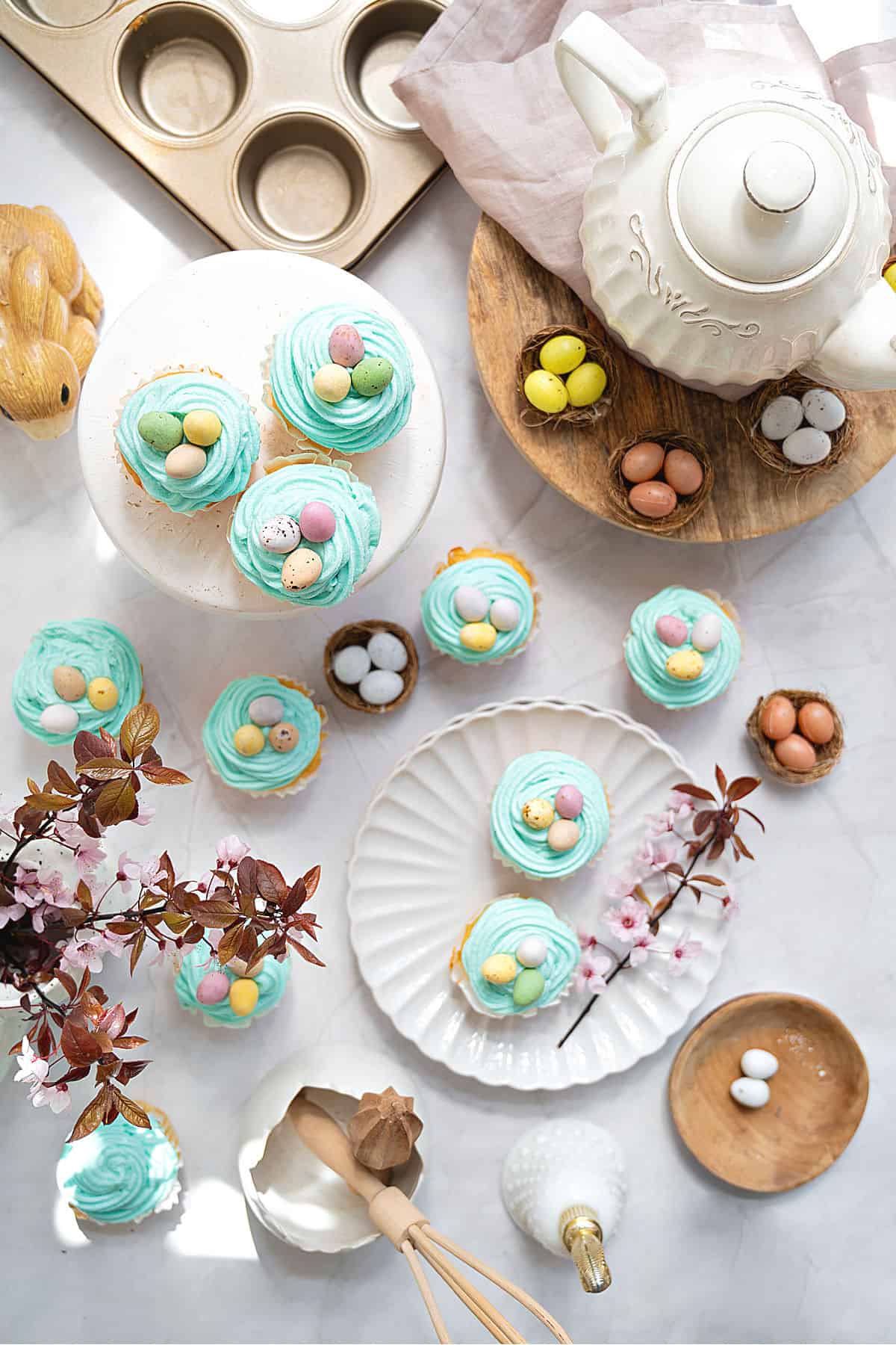 Frosted Easter cupcakes on white plates, teapot, mini eggs. Marbled surface.