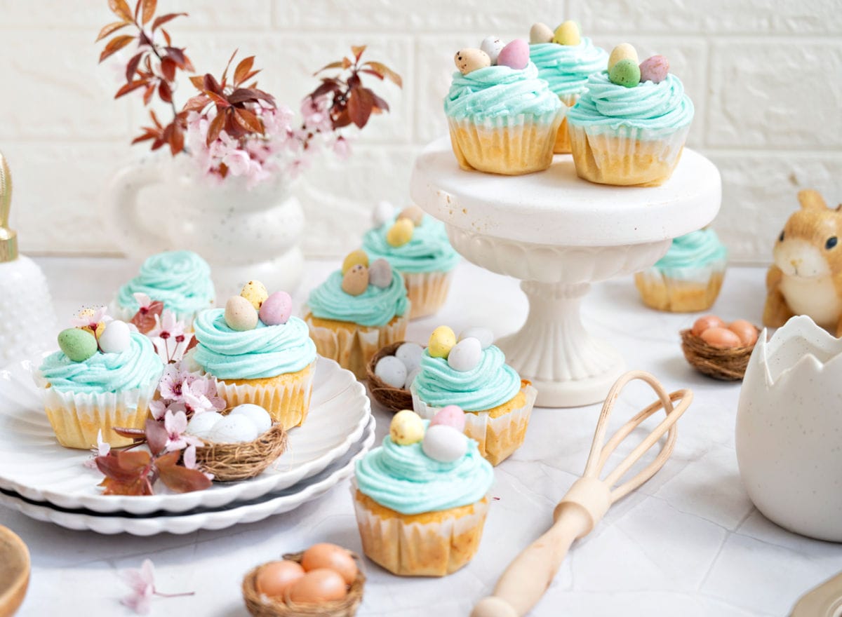 White plate and cake stand with aqua colored Easter cupcakes. Mini eggs, wooden whisk, white background.