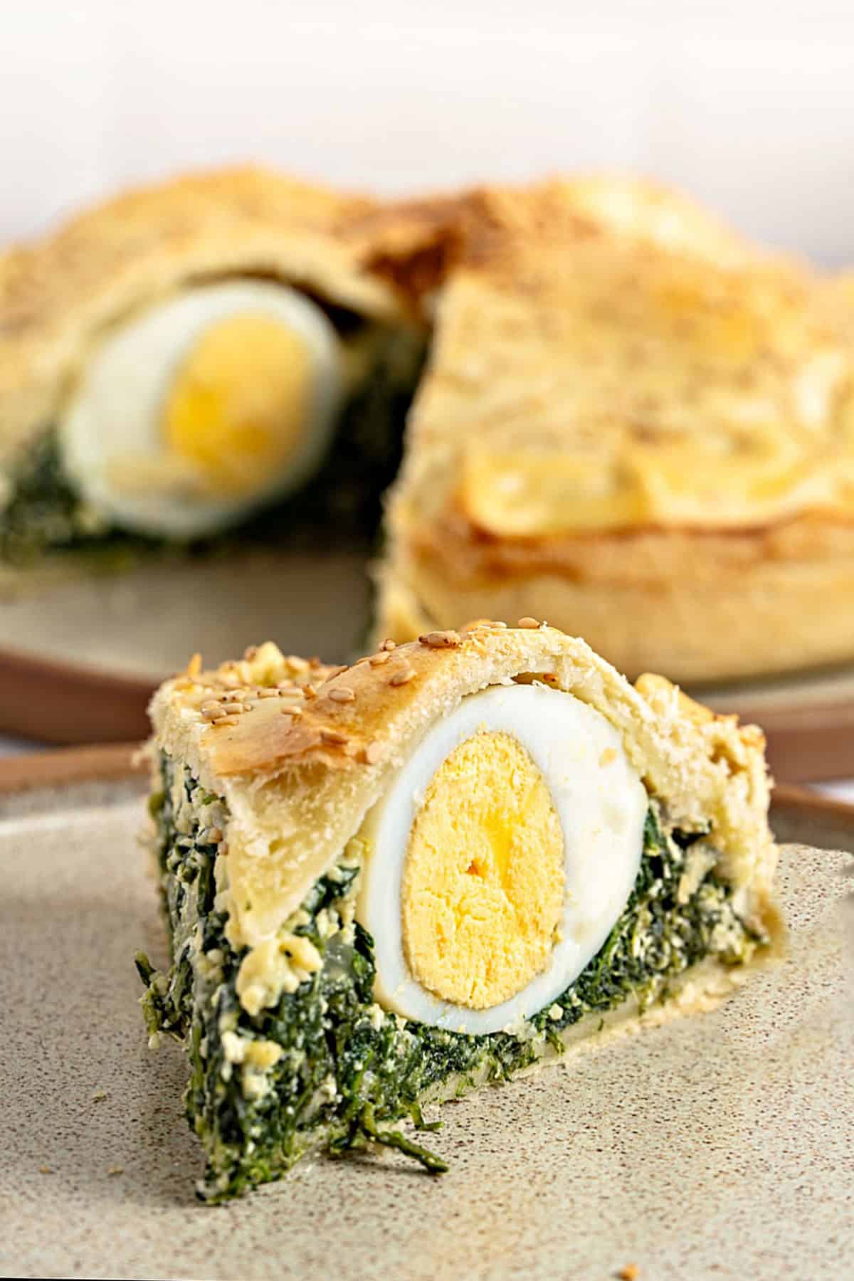 Double crust spinach, hard-boiled egg slice on a beige plate. Whole pie in the background.