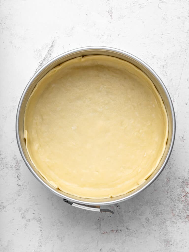 Round springform cake pan lined with pie dough. Light gray background.