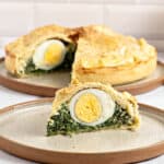 Slice of spinach ricotta pie with hard-boiled egg on a beige plate. Whole tart in the background.