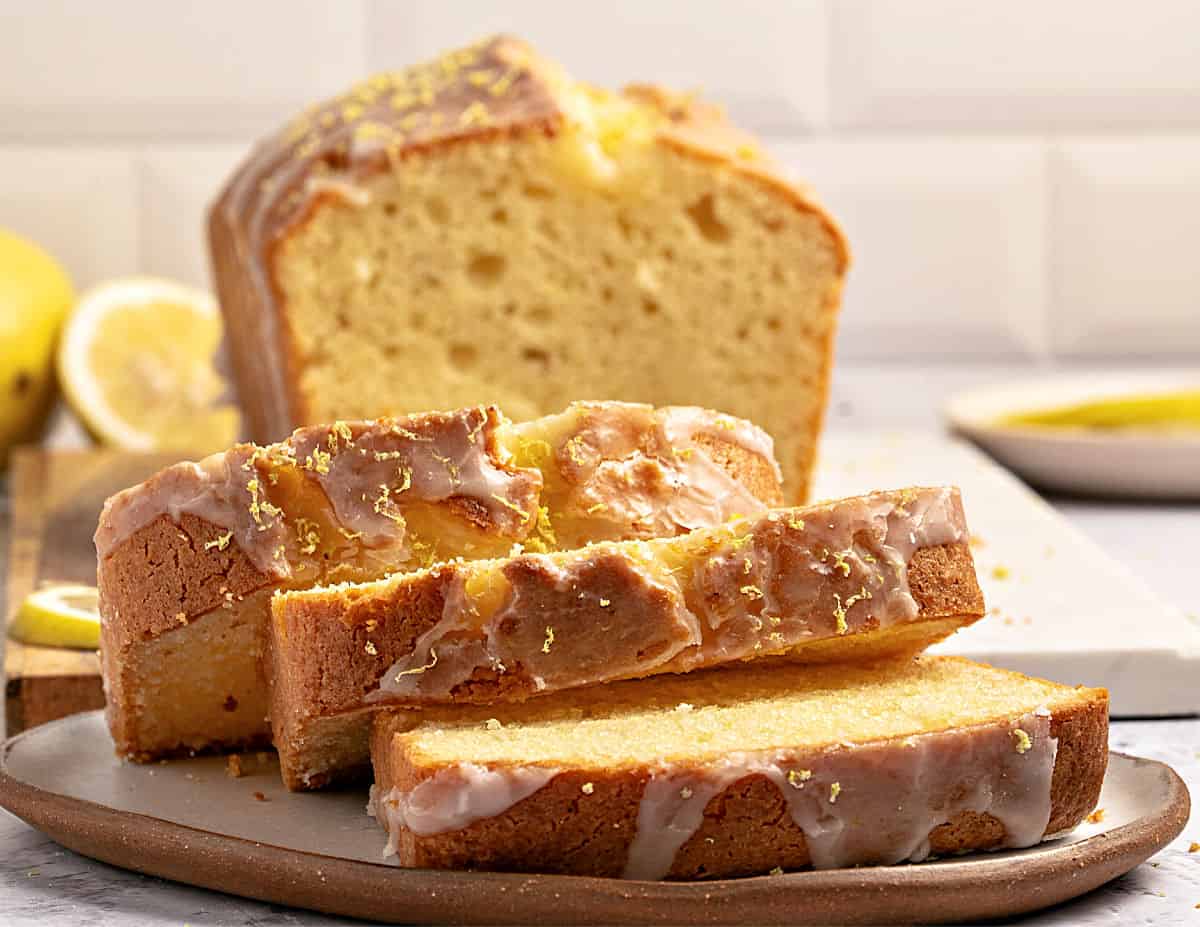 Slices of glazed limoncello loaf cake on a beige plate. White tile background.