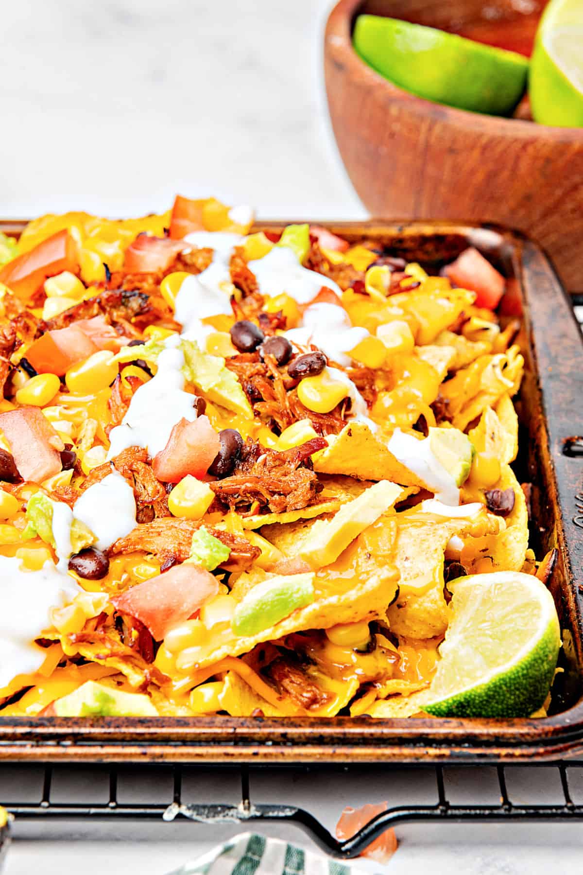 Baking tray with pork, corn, cheese nachos. Sauce drizzle, wire rack, lime wedges.