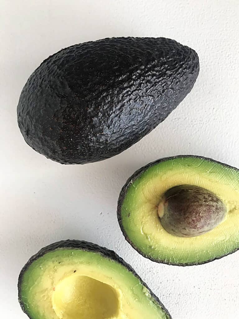 One whole avocado and two halved parts on a white surface.
