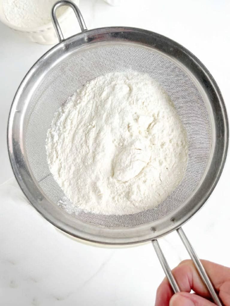 Sifting flour mixture over bowl with cake batter. White surface. Top view.