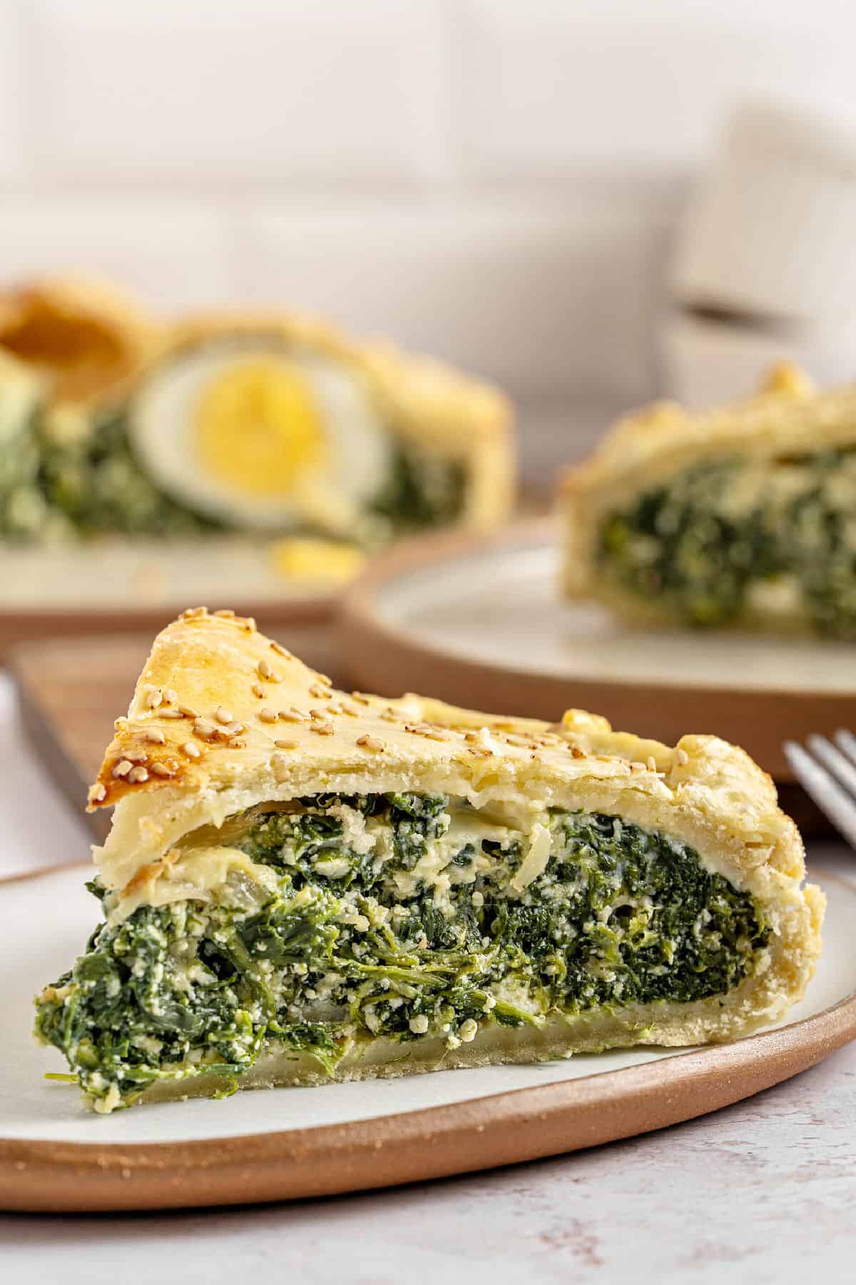 Spinach pasqualina pie slice on a beige plate. More slices on the white tile background.
