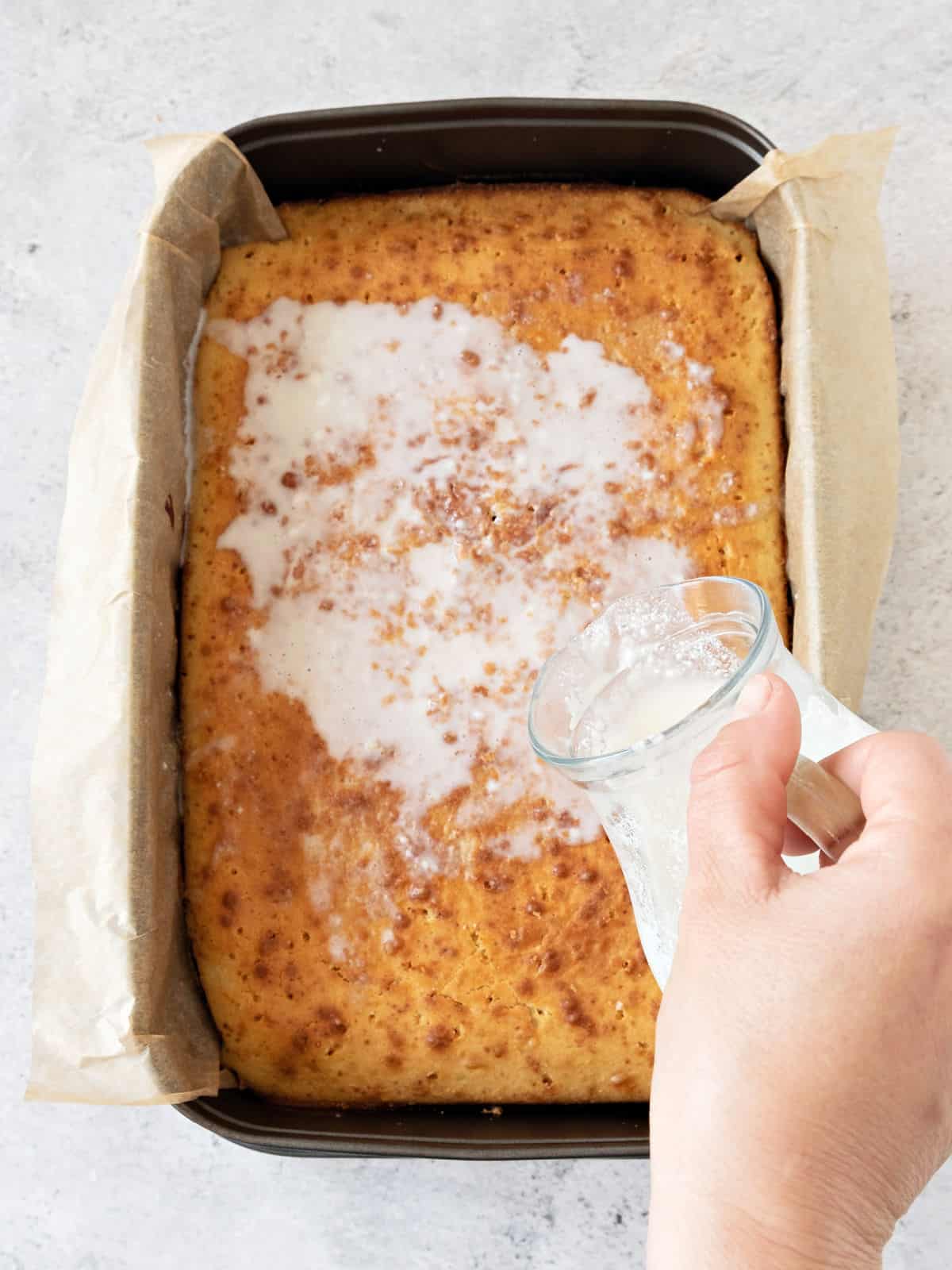 Pouring tres leches mixture on rectangular cake on a dark pan with parchment. Gray surface. Top view.