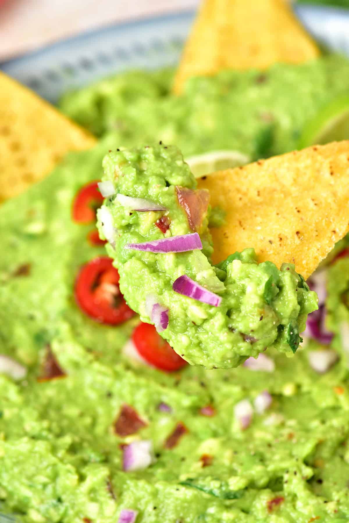 Tortilla chip with guacamole over bowl with more dip. Red onion bits.