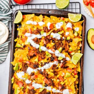 Partial view of metal tray with pulled pork nacho bake, corn, sauce drizzle, lime wedges.