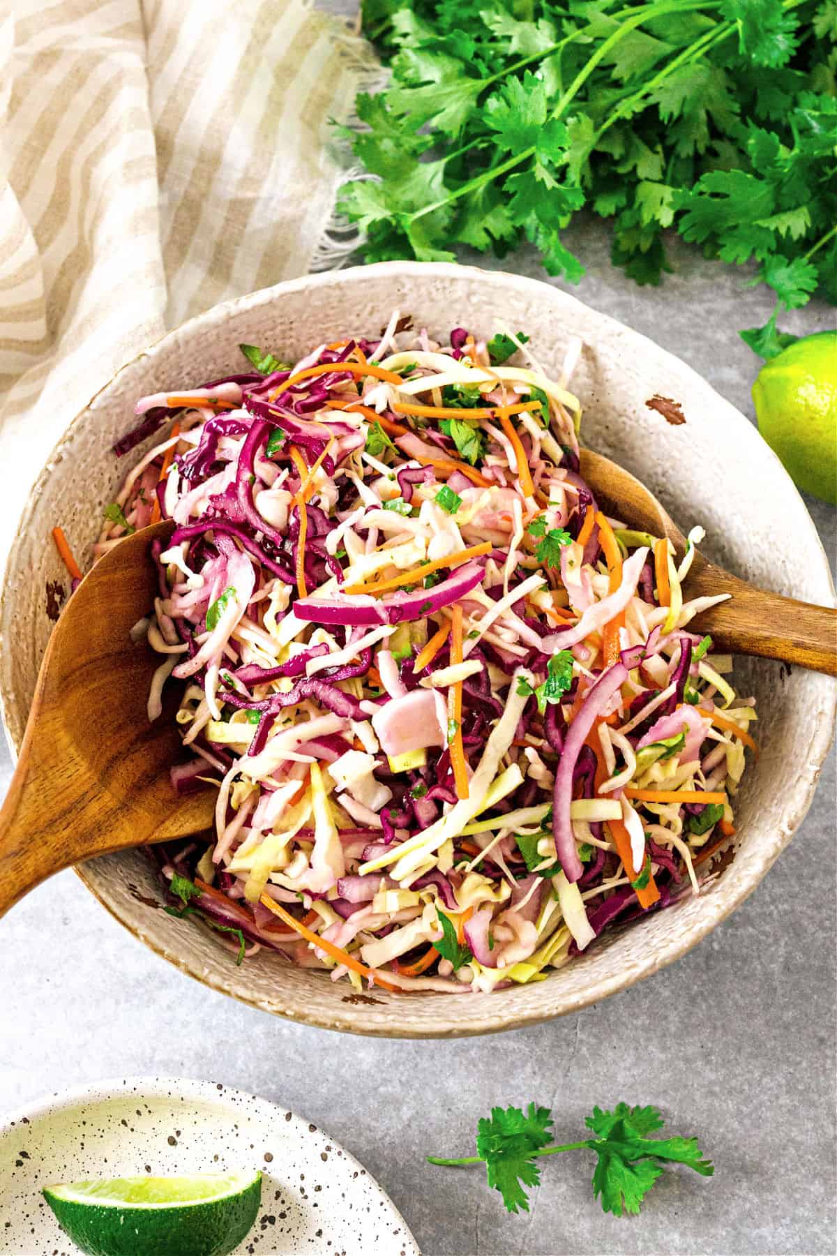 Tossed coleslaw in a white bowl. Wooden utensils, cilantro. Gray background.