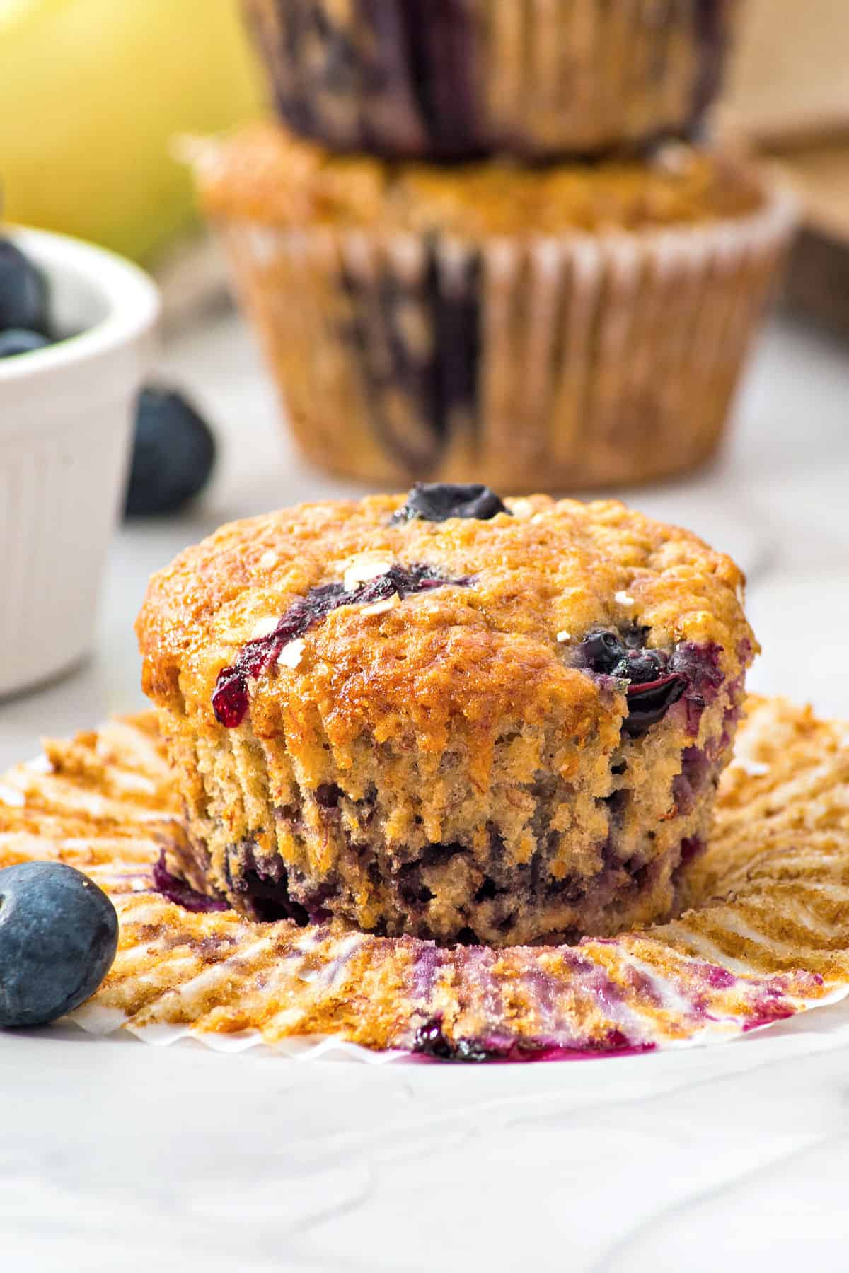 Opened paper liner with oat muffin with blueberries and banana. Muffins and bananas in the background.