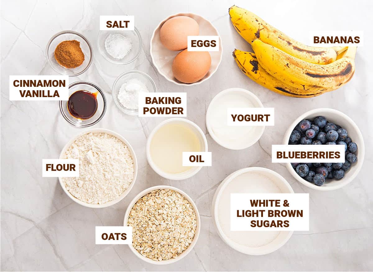 Marbled background with white bowls containing ingredients for banana blueberry oat muffins with oil.