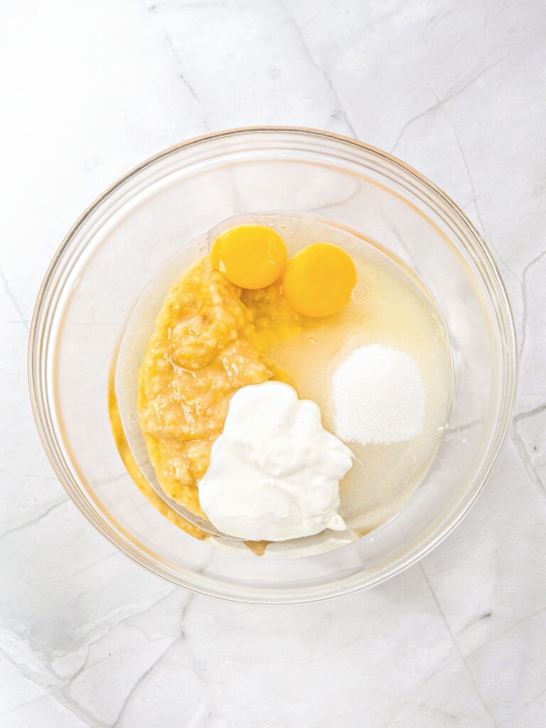 Wet muffin ingredients before mixing in a glass bowl. White marbled surface, top view.