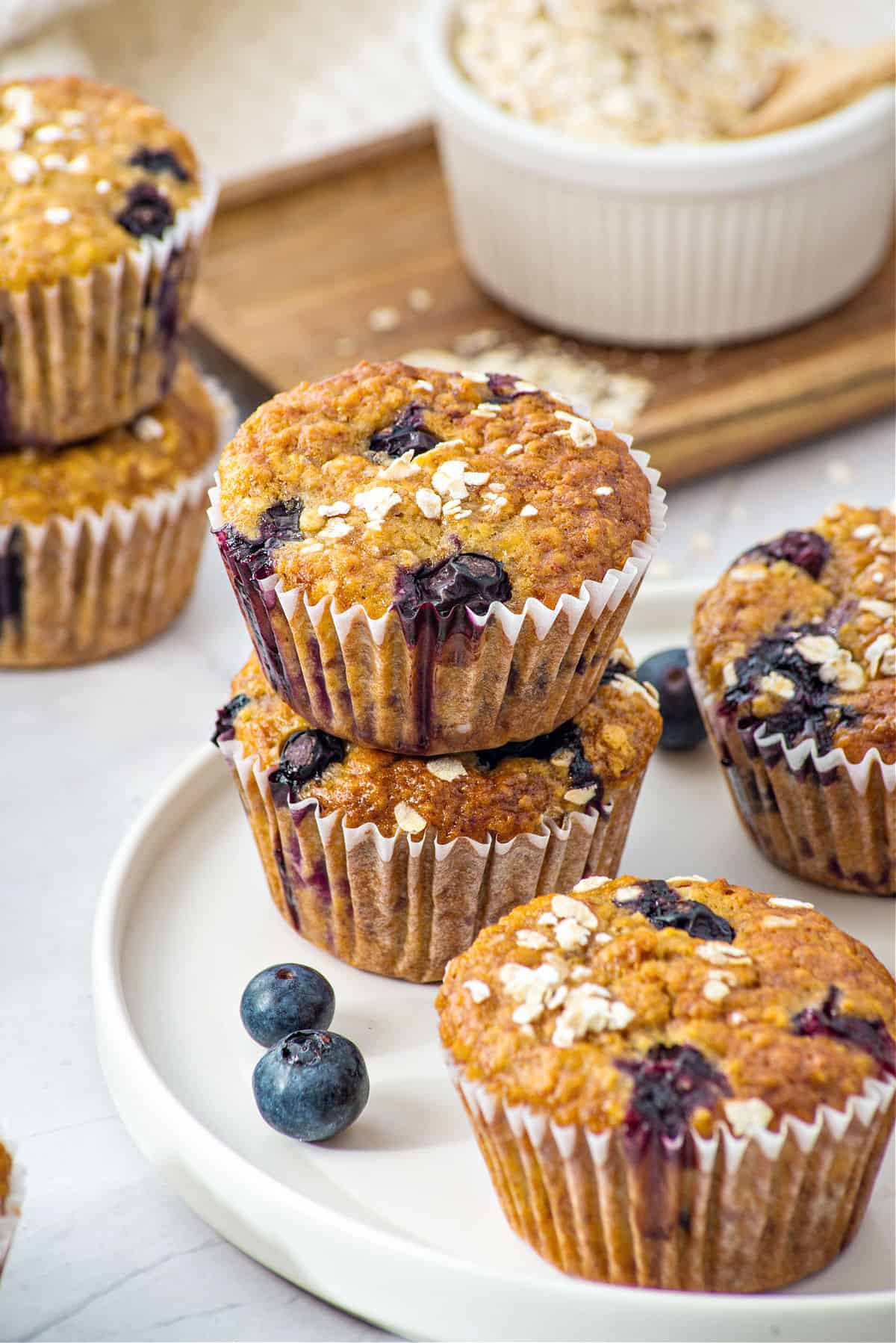 Several blueberry oat muffins on a white plate in paper liners. Fresh berries, bowl with oats.