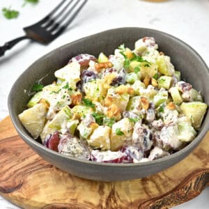 Waldorf salad in a gray bowl on a wooden board. Black fork, white surface.