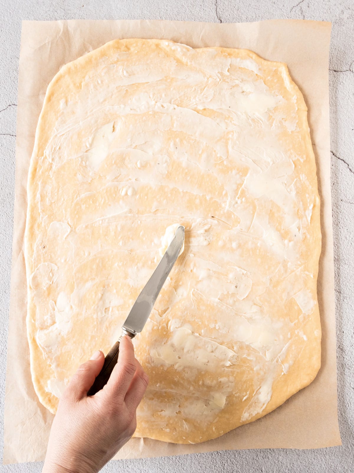 Buttering sheet of cinnamon rolls dough on beige parchment paper.