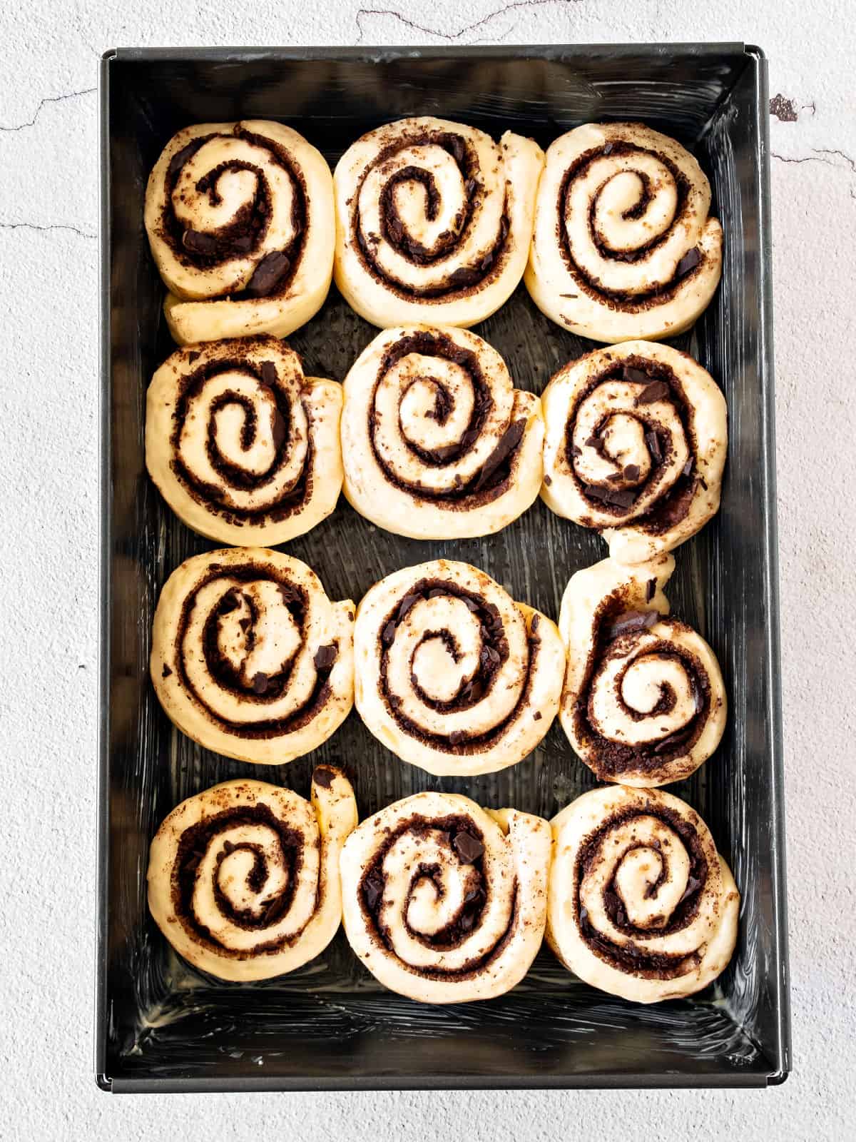 Proofed chocolate cinnamon rolls in a dark baking pan.