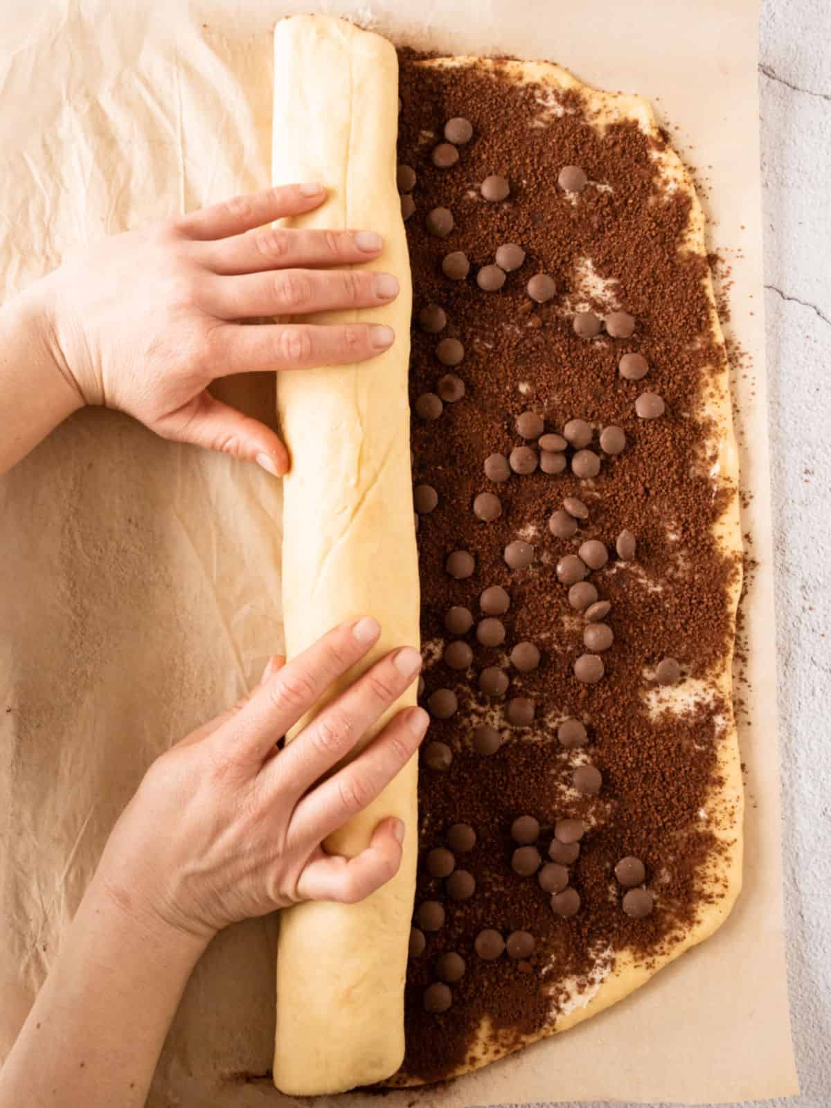 Rolling chocolate filled rolls dough on beige parchment paper.