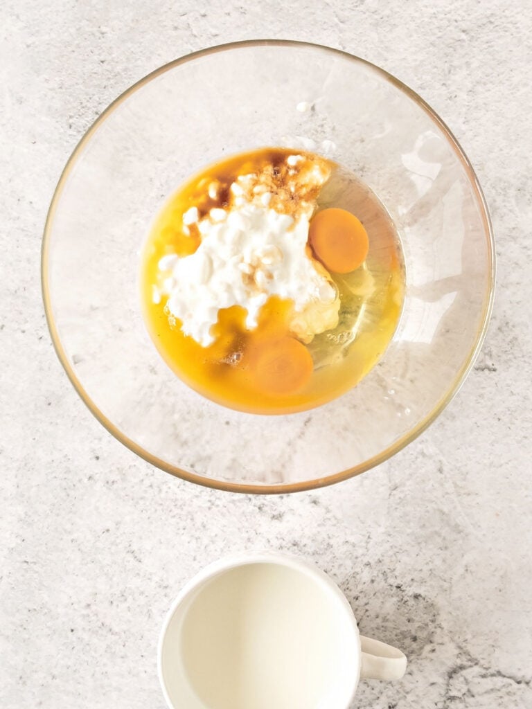 Glass bowl and cup with wet ingredients for cottage cheese pancakes. Light gray background, top view.