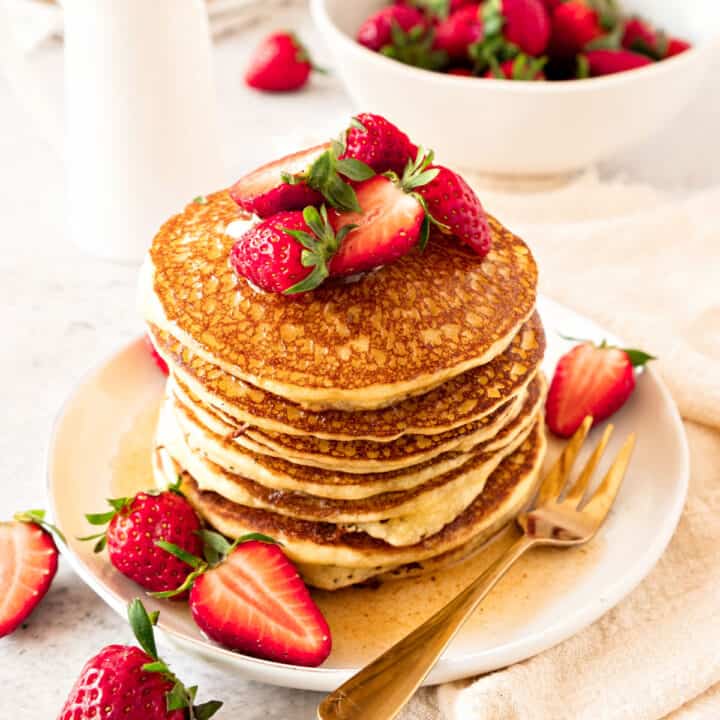 White plate with stack of cottage pancakes with strawberries and syrup. Golden fork, white background.