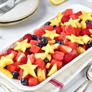 Kitchen towel below white rectangular dish with red, blue and star fruit salad. White background.