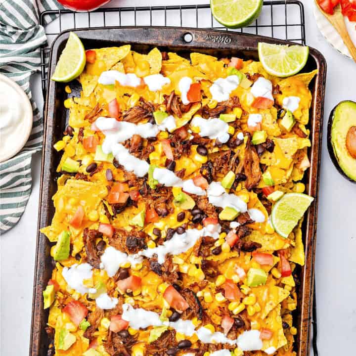 Partial view of metal tray with pulled pork nacho bake, corn, sauce drizzle, lime wedges.