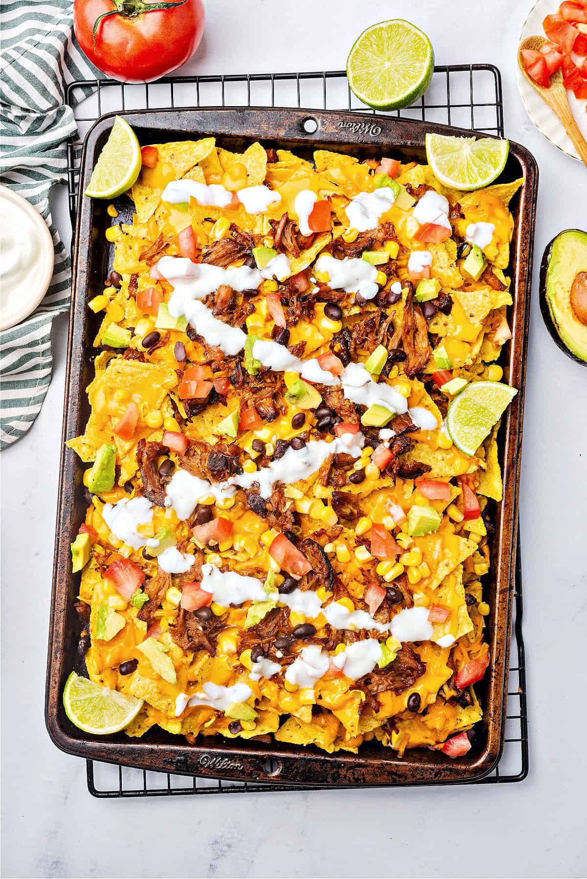 Metal tray with pulled pork nacho bake, drizzle of mayo, lime wedges, tomatoes.
