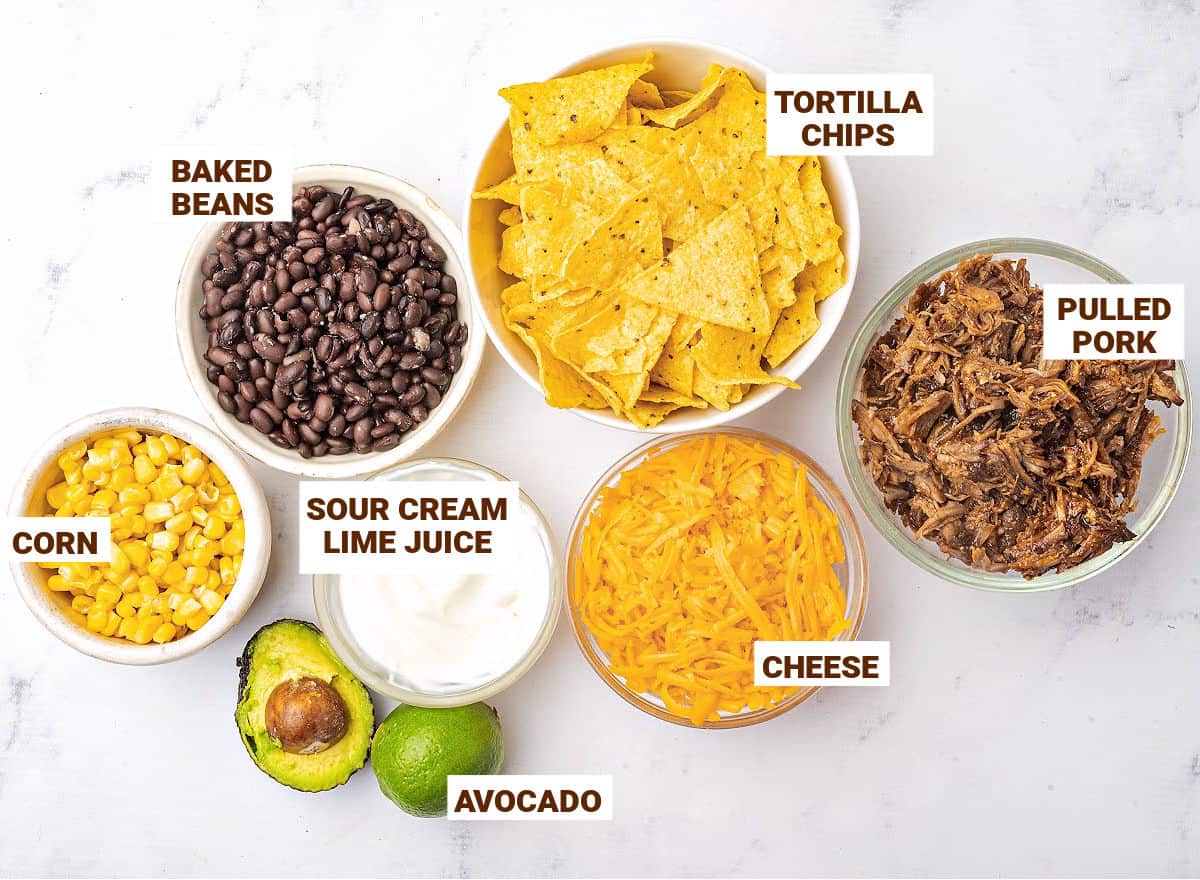 White bowls with ingredients for pork nachos with beans, avocado, corn and sour cream.