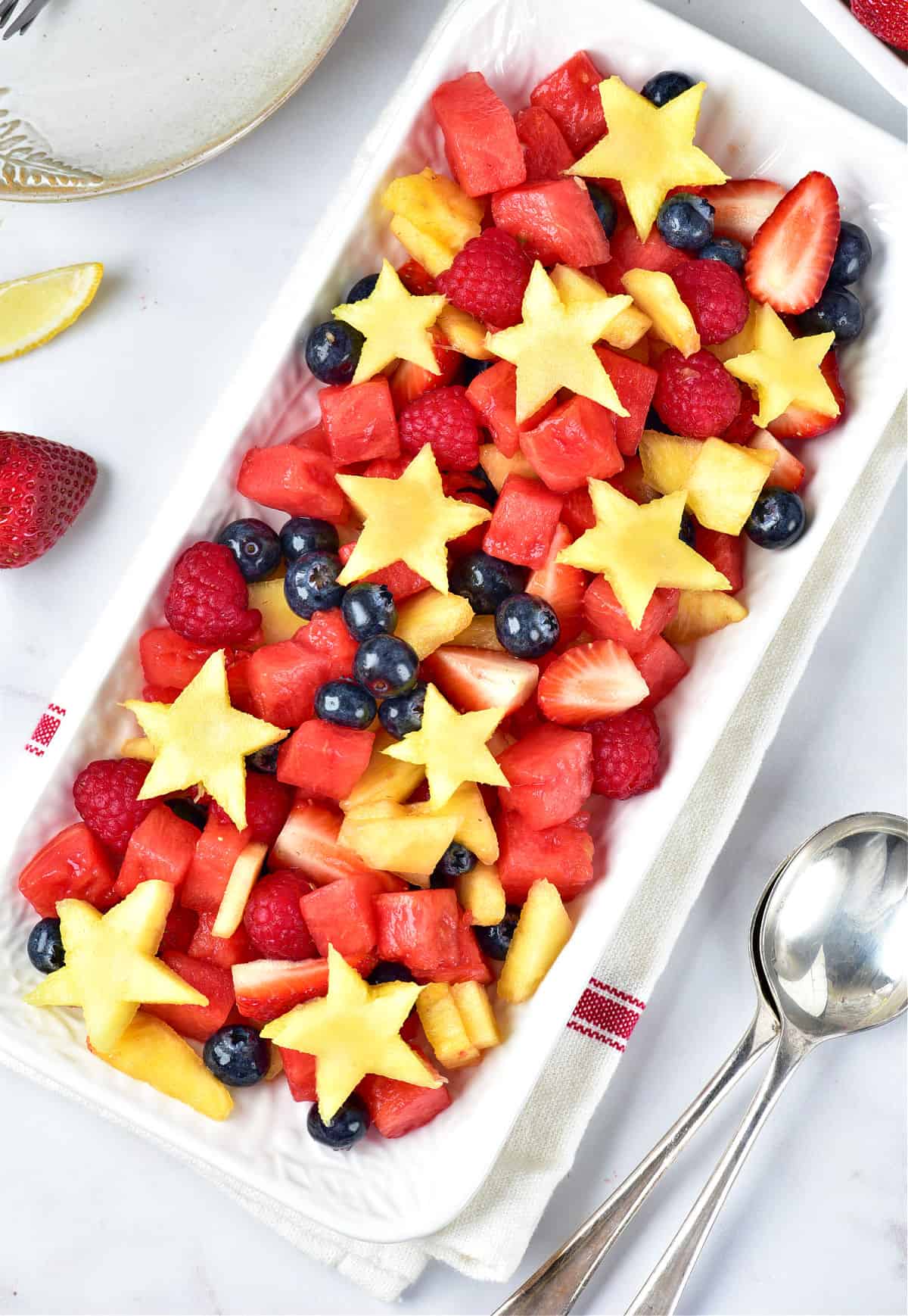 Top view of white dish and surface with patriotic fruit salad. Silver spoons.
