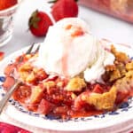 Ice cream topped serving of rhubarb strawberry crisp on a white and blue plate.