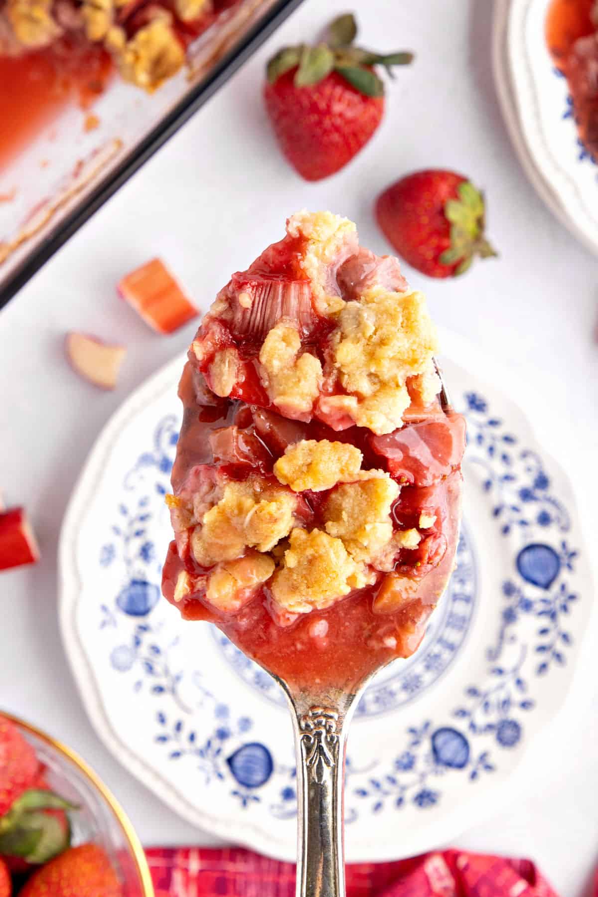 Silver spoon with strawberry rhubarb crisp. White blue plate and berries below.