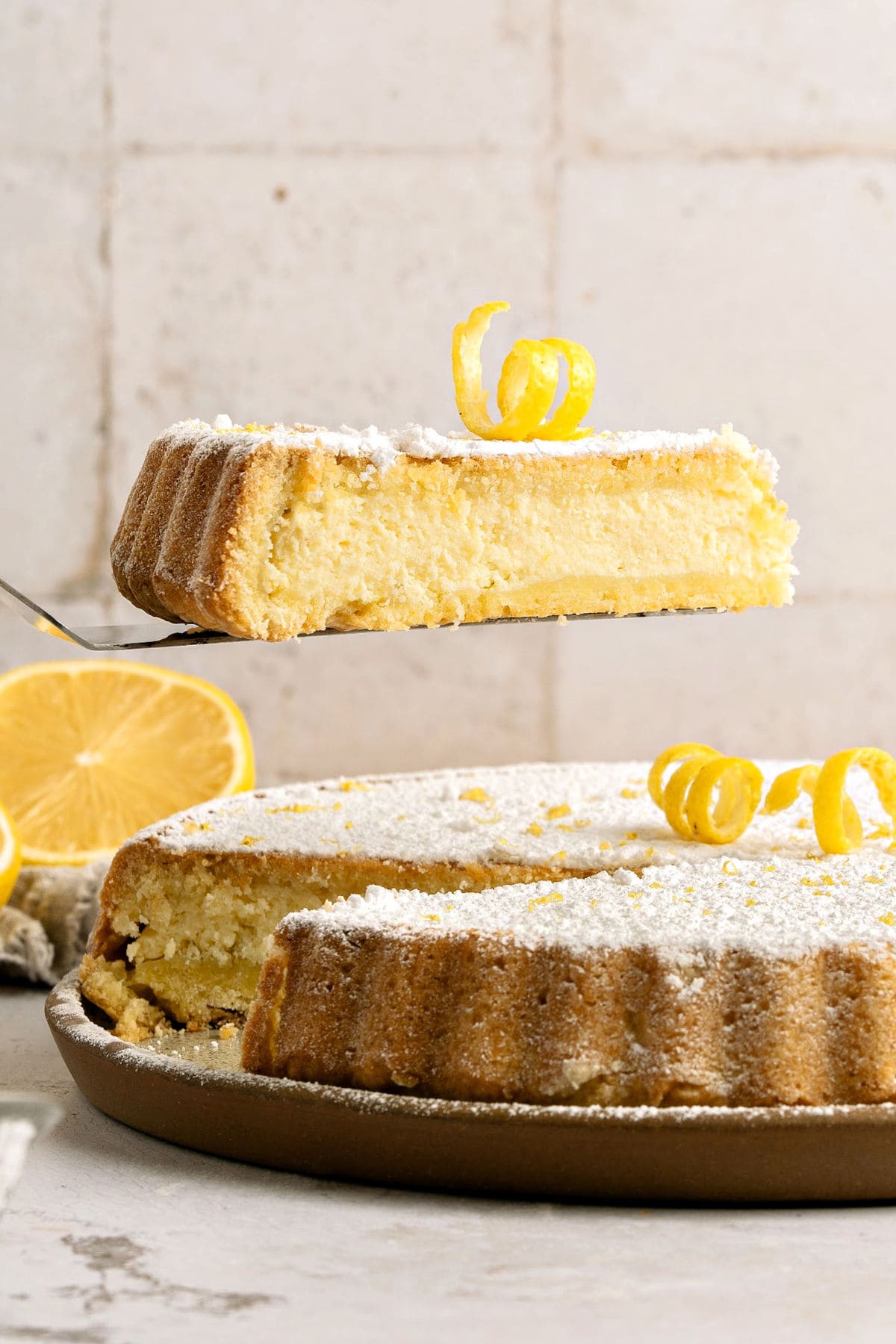 Lifting slice of powdered sugar sprinkled ricotta pie over whole pie on a beige plate. Tile background.