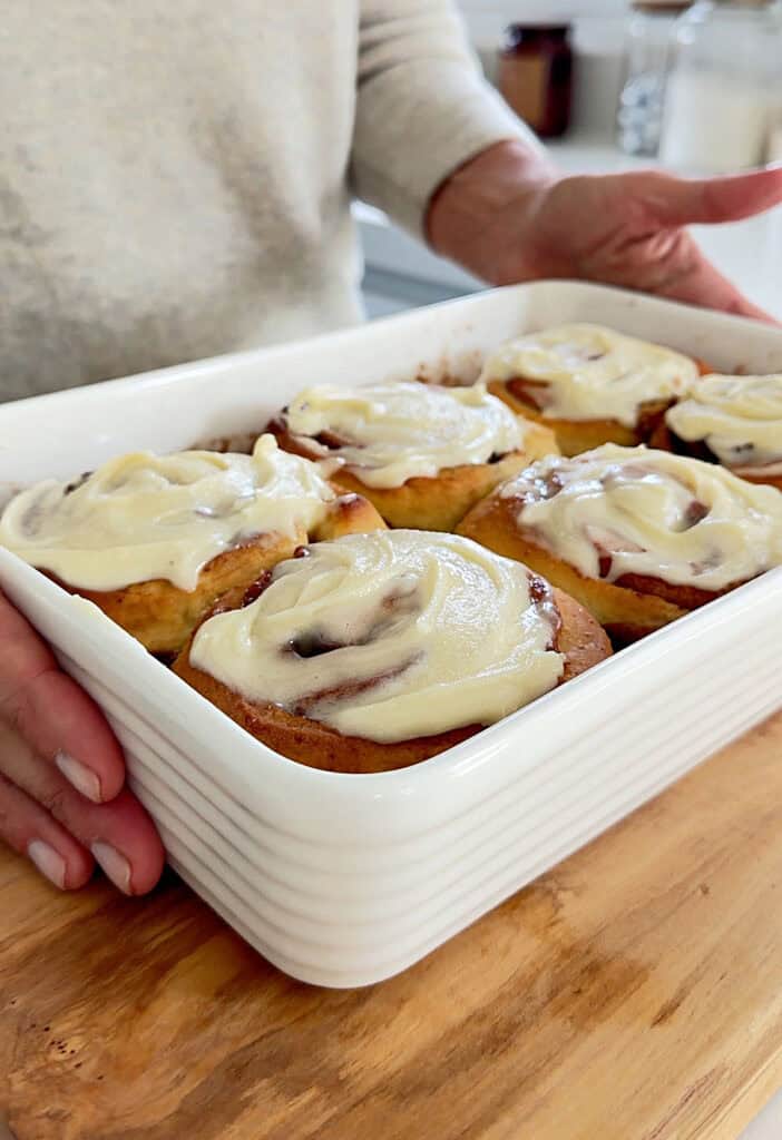 Holding a white baking dish of cream cheese-topped cinnamon rolls on a wooden board.