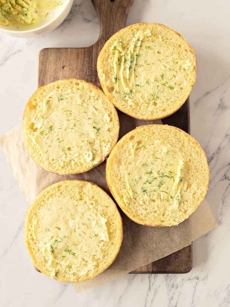 Four burger buns with herb butter on a wooden board. White marble surface. 