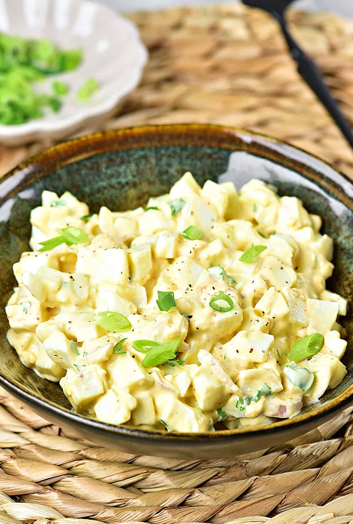 Dark green bowl with egg salad on a woven plate placer.