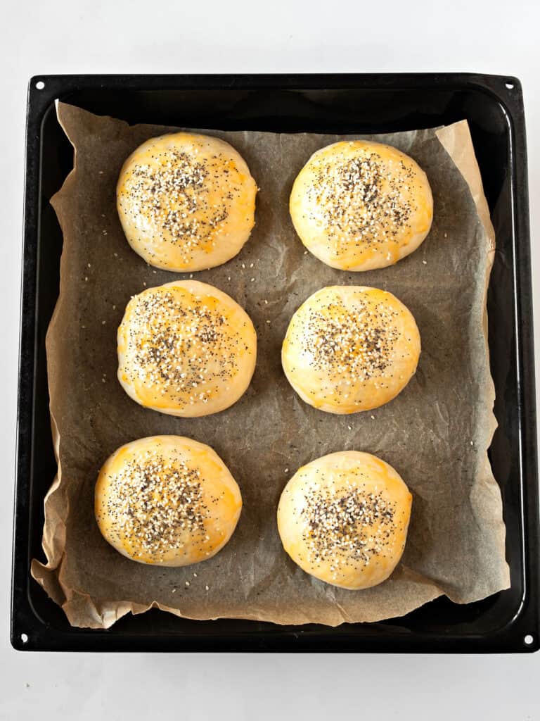 Burger buns with seeds before baking on parchment paper. Black baking tray.